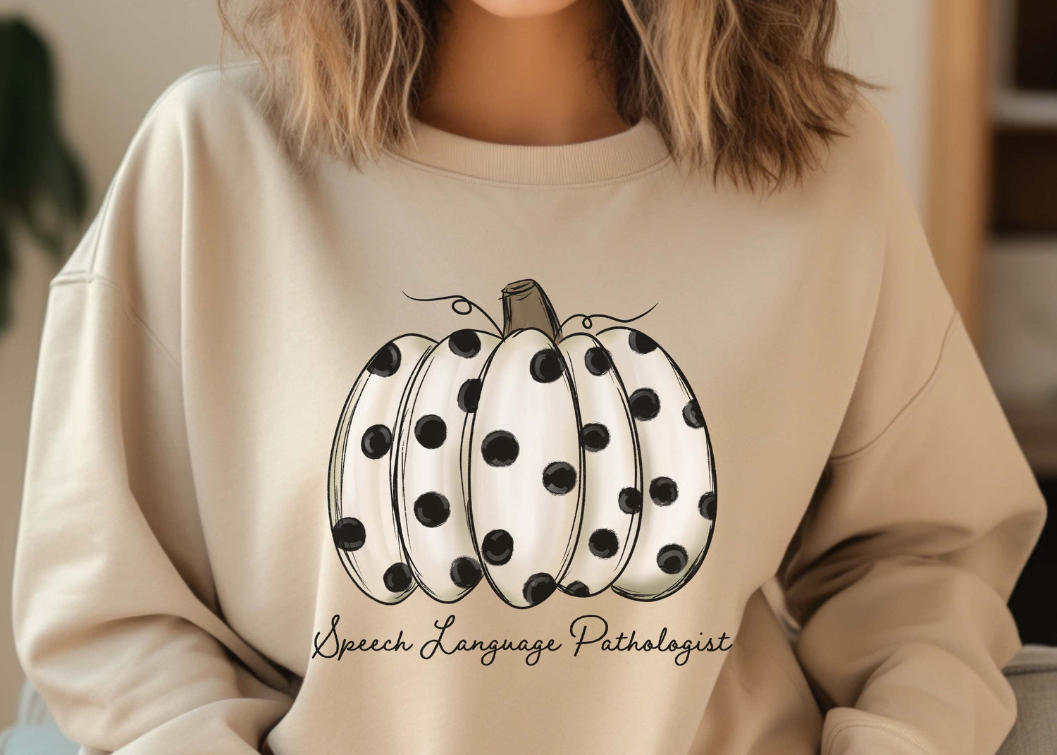 A woman wearing a beige crewneck sweatshirt with a graphic of a white pumpkin with black polka dots and the text 'Speech Language Pathologist' written below it.