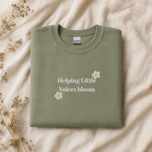A folded sage green crewneck sweatshirt with the text 'Helping Little Voices bloom' and small daisy graphics on the front, displayed on a beige fabric background with dried flowers.