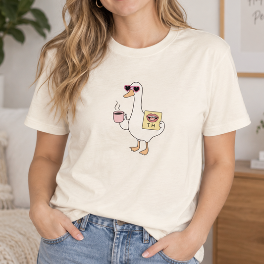 A woman wearing a cream-colored graphic t-shirt with a cartoon duck wearing sunglasses and holding a coffee cup and a bag of coffee, paired with blue jeans in a cozy, modern living room setting.