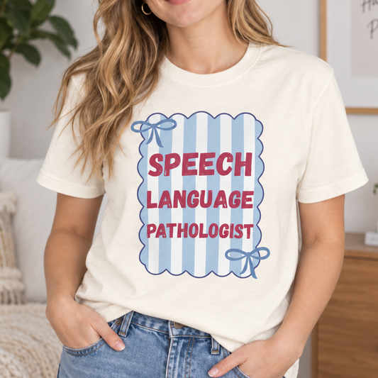 A woman wearing a cream-colored graphic t-shirt with a blue and red striped design that reads 'SPEECH LANGUAGE PATHOLOGIST' in red letters, paired with blue jeans, standing in a cozy, modern living room setting.